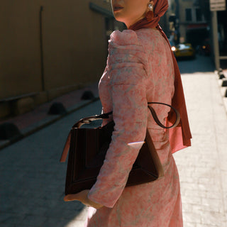 A woman wearing a light pink floral long-sleeve dress and a dusty rose headscarf stands on a sunlit city street, carrying a dark brown leather handbag.